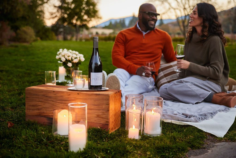 A couple sits on a blanket in the grass, drinking wine and surrounded by candles